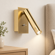 Aplique de lectura dorado con placa rectangular encendido sobre pared clara junto a cabecero y mesilla de madera